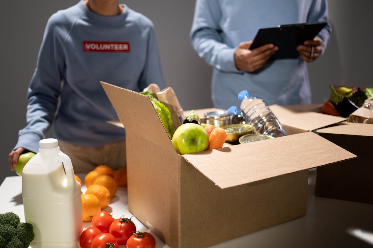 hero-homepage People packing a cardboard box with essentials like fruits, vegetables, and bottled water for charity.